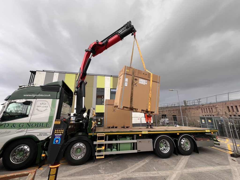 A photograph of a large boxed air source heat pump being crane lifted off a lorry in front of the Stella Turk building at Penryn campus.   