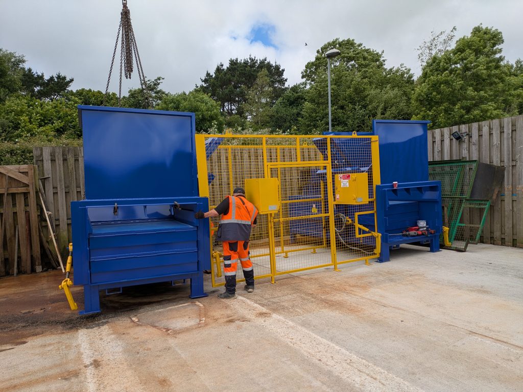 The two new compactors being positioned in the waste compound and a man in hi-visibility clothing directing the lift into position.
