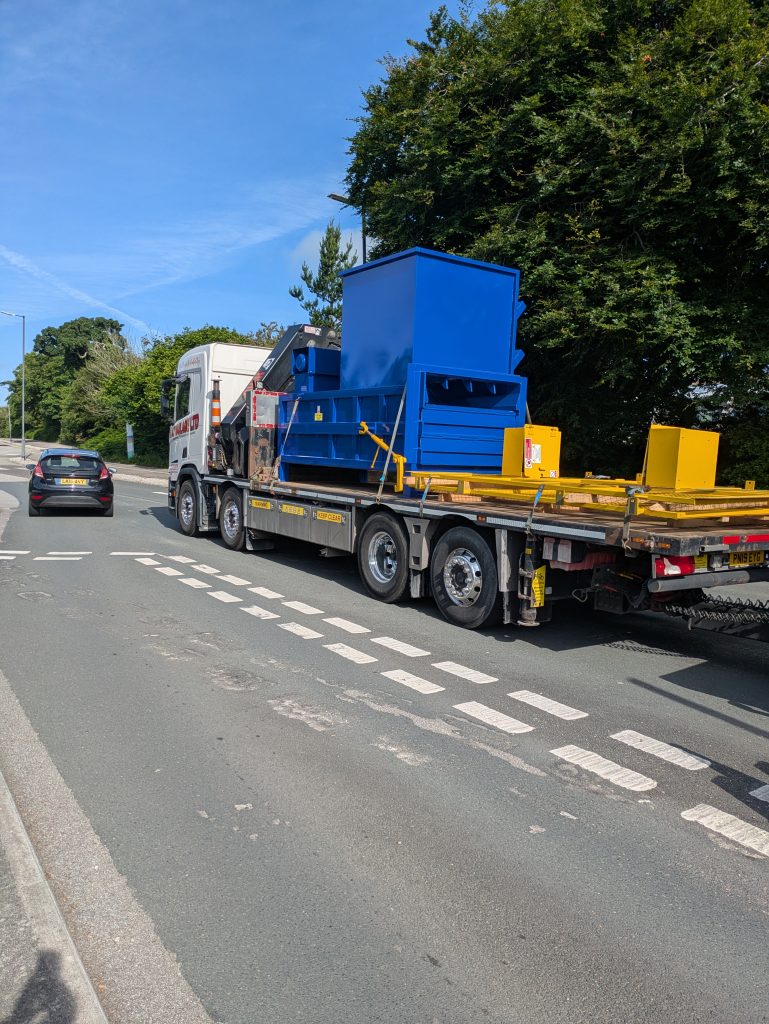 A lorry with lifter transporting two large blue and yellow waste compactors on to trailers.