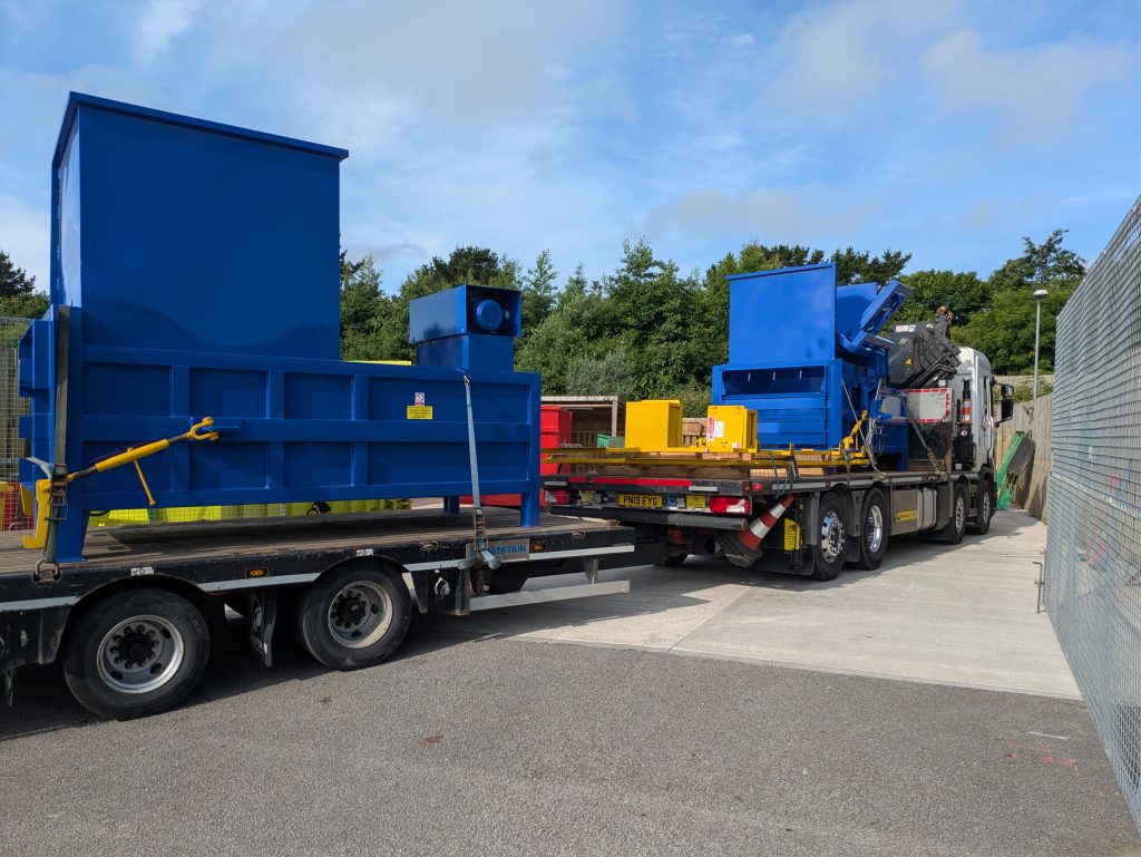Two blue metal waste compactors pictured on the two trailers being pulled by a large lorry.