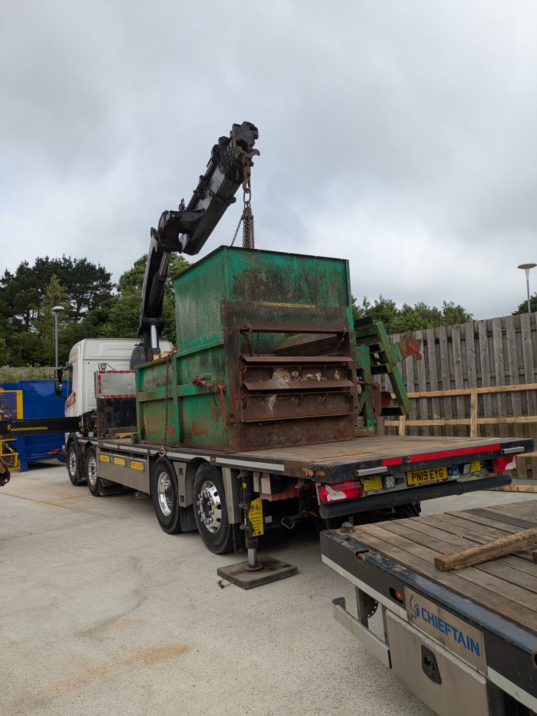 A lorry with lifter lifts a large, rusting metal waste compactor onto a trailer to take it away.