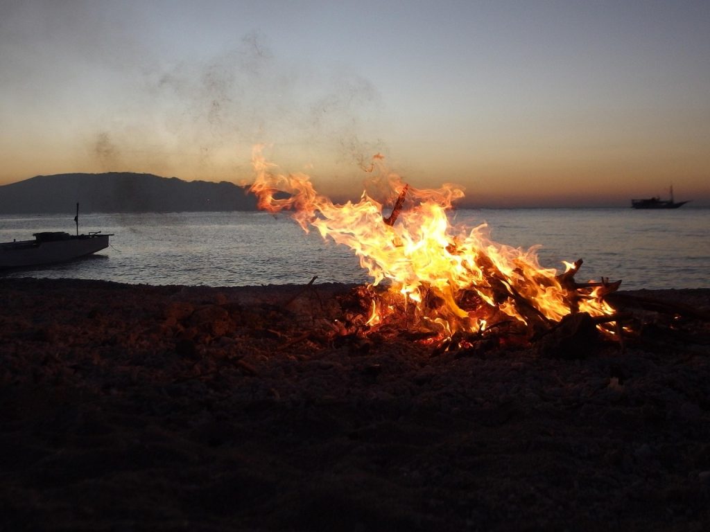 Image of a bonfire on a beach, looking out across the ocean with the silhouette of bots in the background